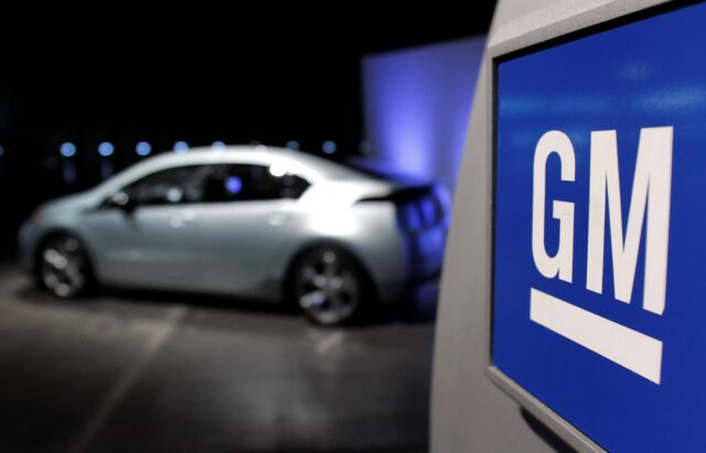 A sign reading "GM" with a Chevy Volt blurry in the background